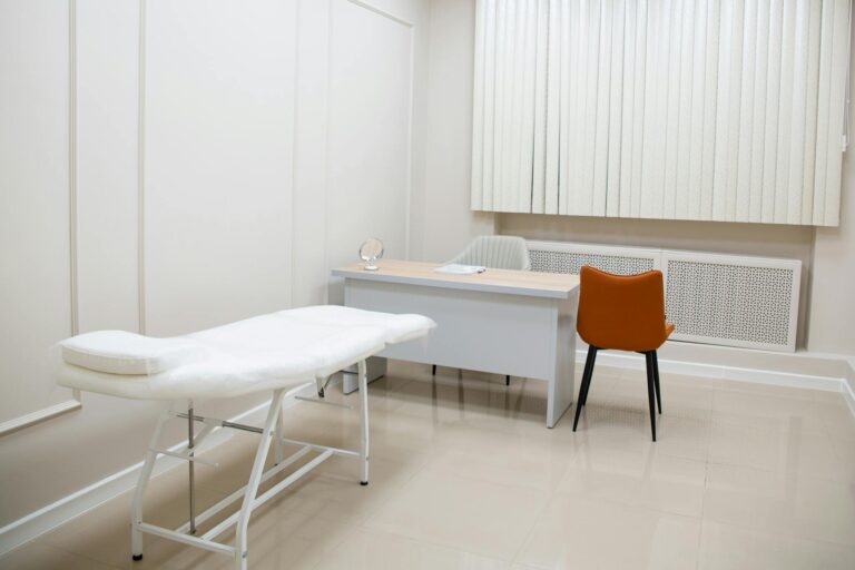 A minimalist medical examination room featuring a desk, chairs, and an examination bed in a sterile, neutral setting.