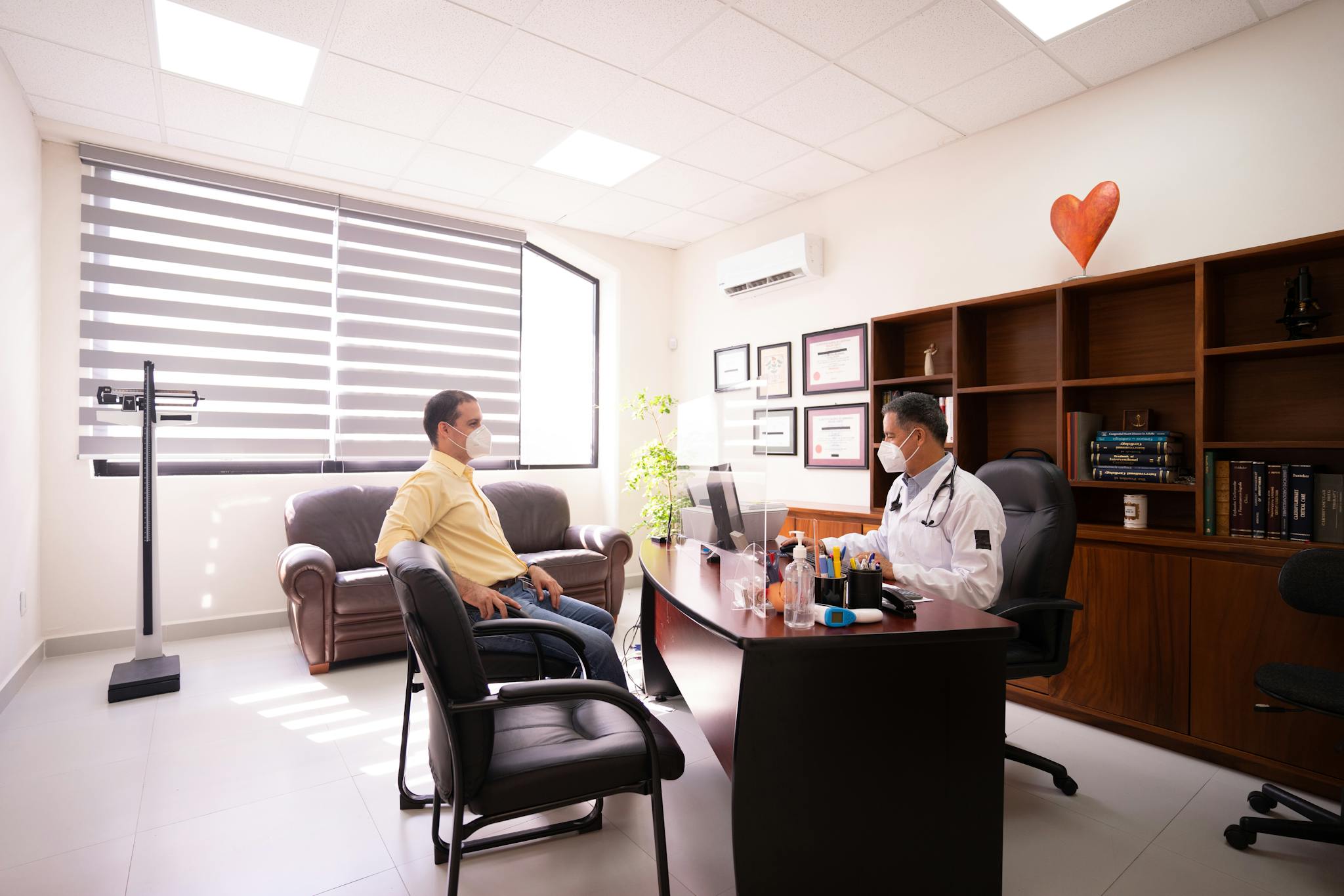 A patient consults with a masked doctor in a well-lit, modern office. Safety measures in place.